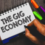 Word writing text The Gig Economy. Business concept for Market of Short-term contracts freelance work temporary Hand holding pen and paper sketch words near lie some pen on woody desk.
