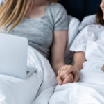 partial view of two lesbians with laptop and book holding hands while lying under blanket in bed