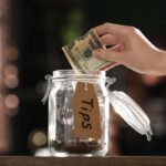 Woman putting tips into glass jar on wooden table indoors, close