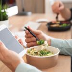 Office employee with smartphone having lunch at workplace, closeup. Food delivery